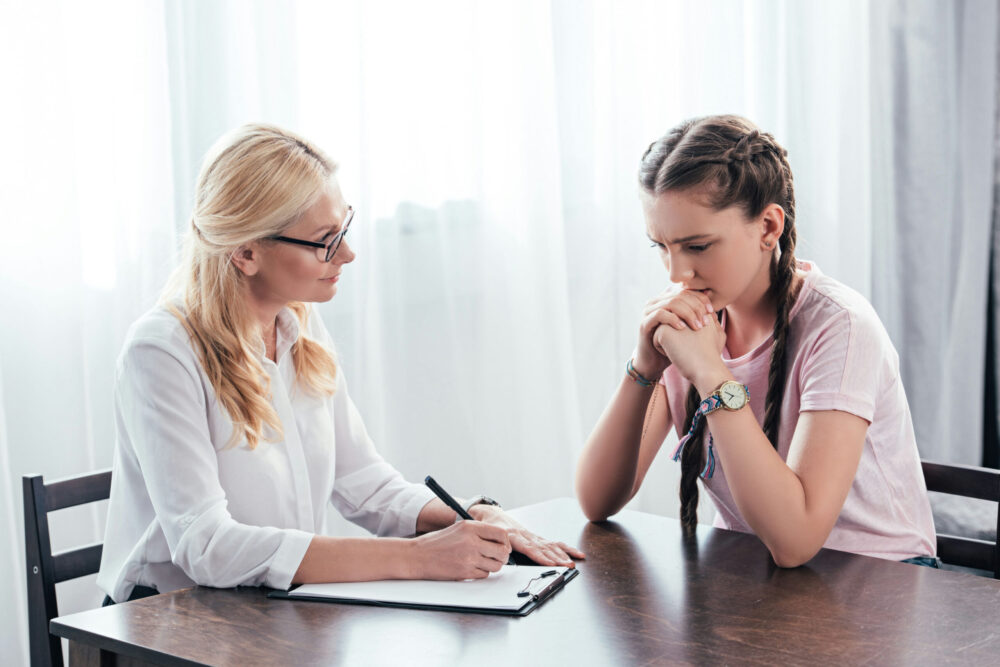 worried-teen-girl-at-table-with-blonde-female-counselor-taking-notes